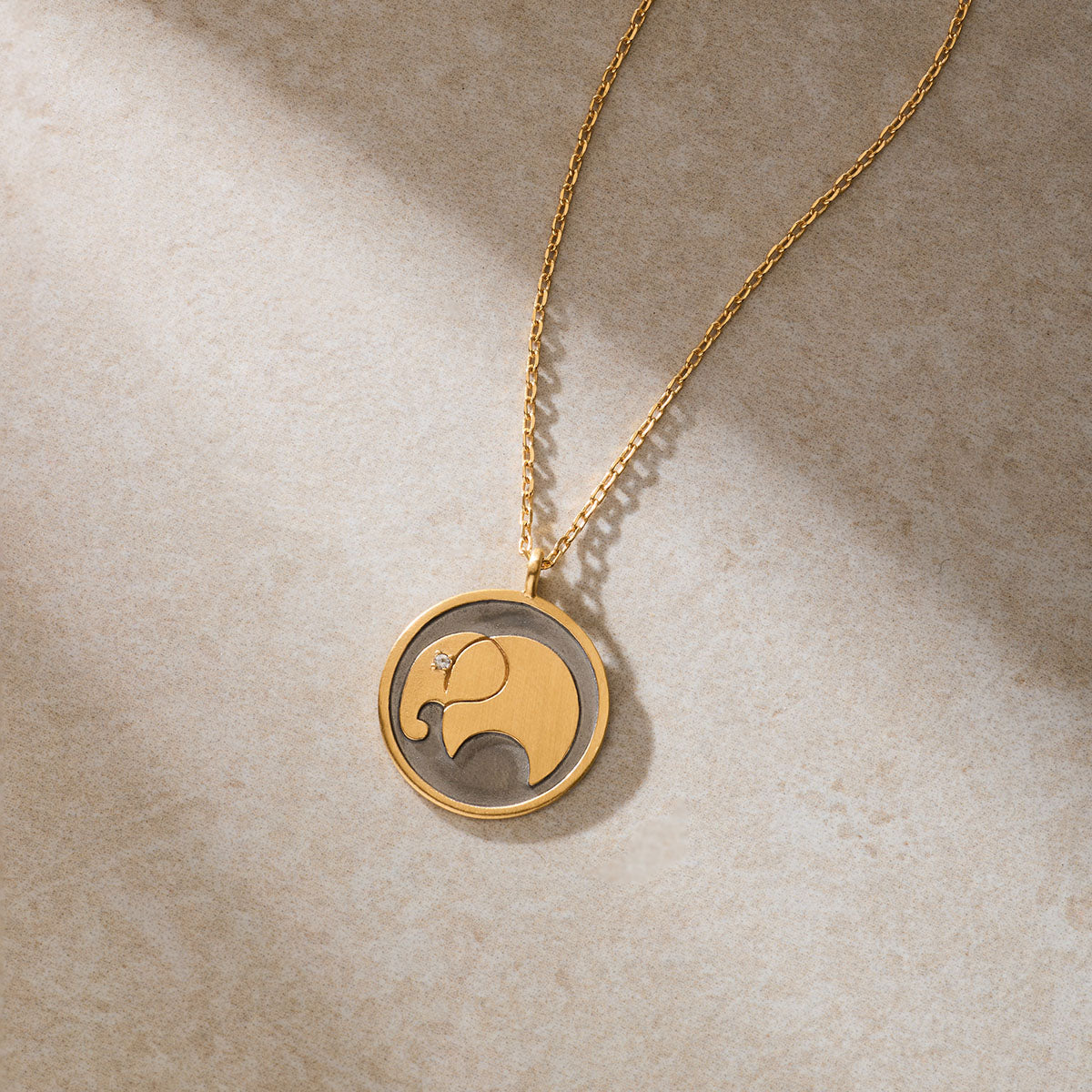 A gold chain necklace with a round pendant featuring a stylized gold elephant with a small gemstone eye rests on beige stone.