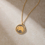 A gold chain necklace with a round pendant featuring a stylized gold elephant with a small gemstone eye rests on beige stone.