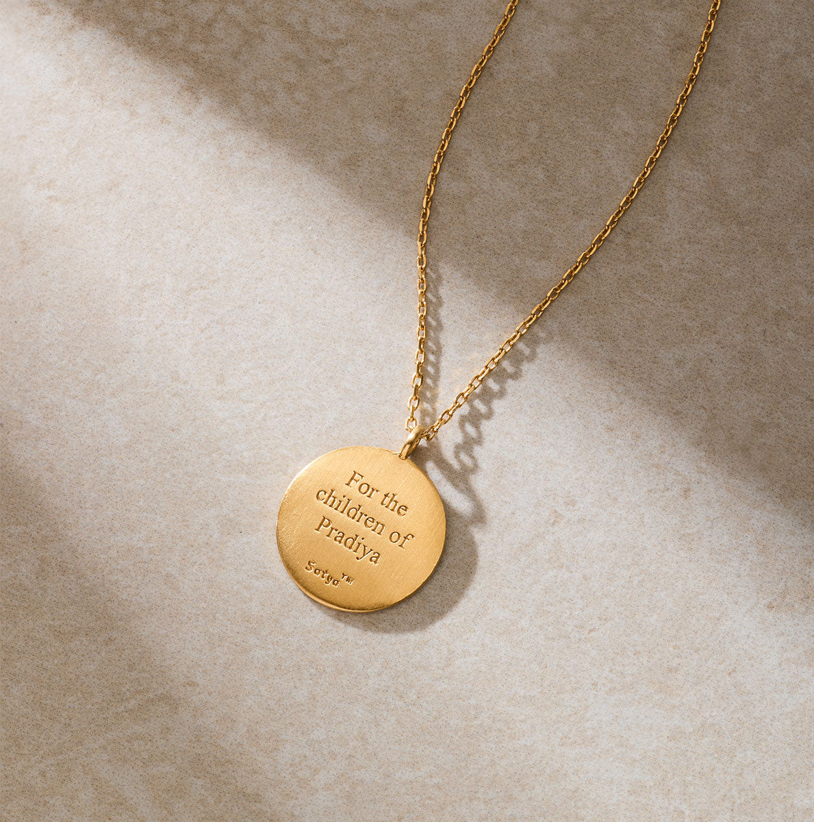 A gold necklace with a round pendant engraved "For the children of Pradiya" rests on a beige stone surface.