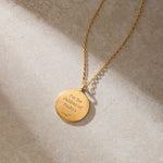 A gold necklace with a round pendant engraved "For the children of Pradiya" rests on a beige stone surface.