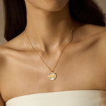 A woman wears a thin gold chain necklace with a round pendant featuring a gold elephant on a pale background.