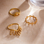 Three gold band rings with decorative details including a sun motif, beaded texture, and dangling leaf charms on a neutral su