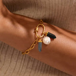 Gold chain bracelet with white pearl charm, black stone bead, and gray tapered stone pendant worn on wrist.