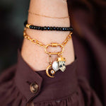 Gold chain bracelet with black beaded strand and hanging charms including a pearl, moon, and spotted stone pendant.