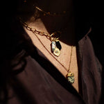 Gold charm pendant with iridescent stone and eye motif on layered chain necklace worn on person.