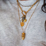 Gold-toned layered necklaces with pearl, sun, and pointed charm pendants worn on light gray fabric.