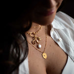 Gold-toned charm necklace with pearl drop and celestial pendants worn on model's chest.