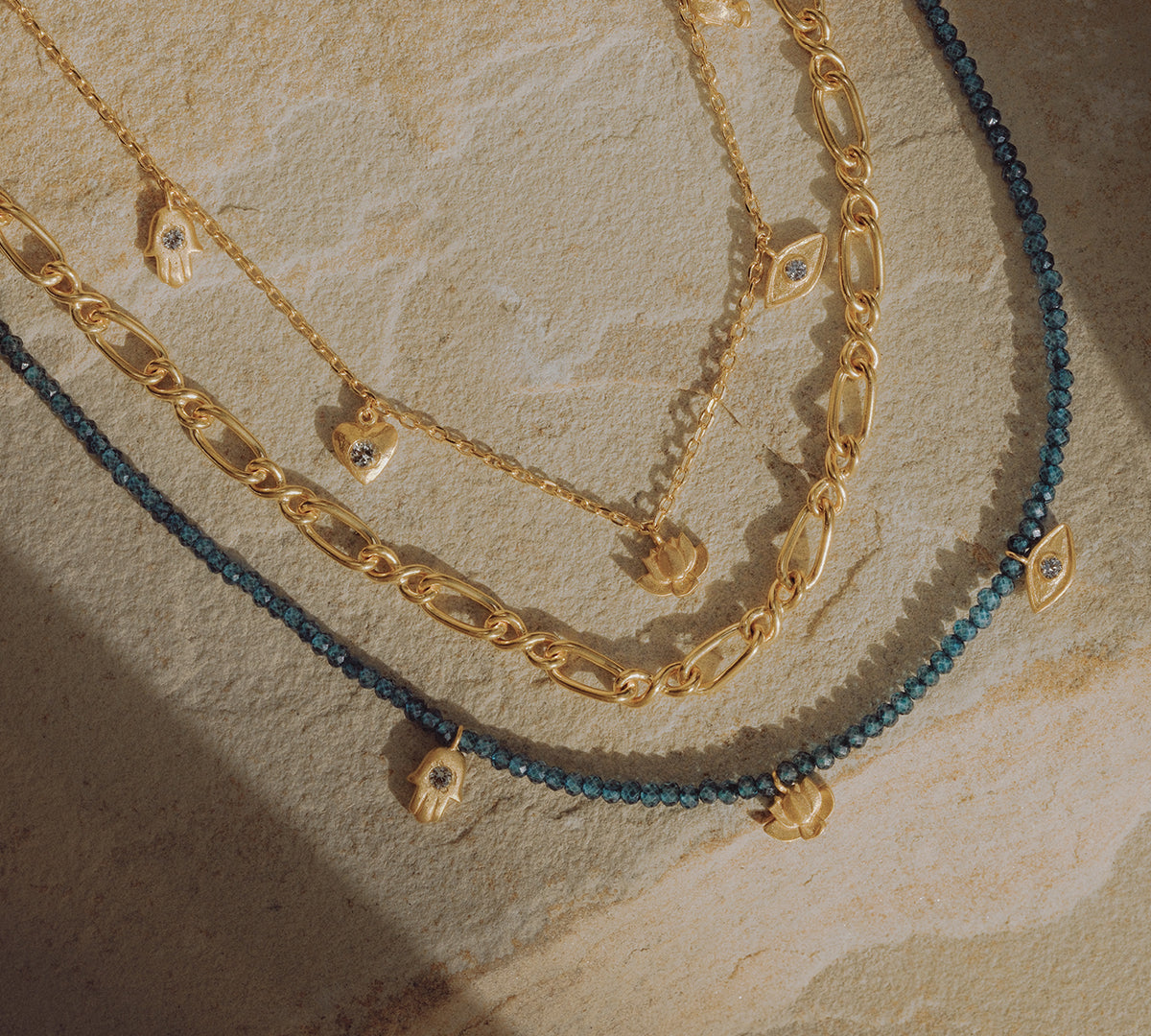 Three layered chain necklaces in gold and blue with small pendant charms on textured background.